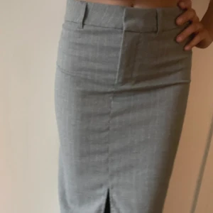 Grå långkjol med slits - Snygg grå långkjol i klassisk modell med hög midja och slits framtill. Kjolen har bälteshällor och en stilren look, perfekt för dig som gillar en clean och enkel stil. Materialet känns som bomull eller polyesterblandning.