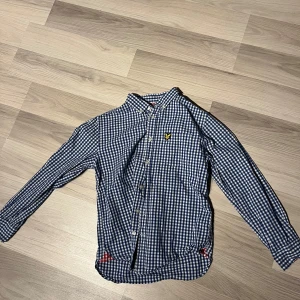 Blårutig skjorta från Lyle & Scott - Snygg blårutig skjorta från Lyle & Scott Junior med klassisk krage och knappar framtill. Skjortan har lång ärm och det ikoniska gula logobroderiet på bröstet. Tillverkad i mjuk bomull och har rödrutigt tyg på insidan av kragen för extra detalj.