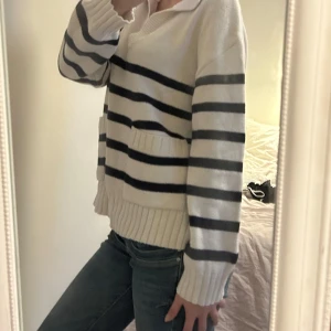 Vit och marinblå randig stickad tröja - Säljer en vit stickad tröja med marinblå ränder och två fickor framtill. Tröjan har en krage med v-ringning och breda ribbade muddar vid ärmslut och nederkant. Perfekt för en chill och snygg look.