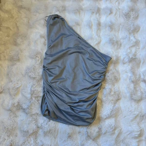 Grå one shoulder topp Gina Tricot - Snygg grå topp från Gina Tricot med one shoulder-design och rynkade detaljer längs sidan. Toppen är tight och tillverkad i mjuk polyesterblandning som sitter skönt mot kroppen. Perfekt för dig som vill sticka ut med en modern och stilren look.