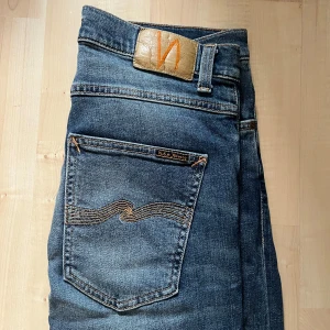 Blå jeans från Nudie Jeans, W31 L34 - Säljer ett par klassiska blå jeans från Nudie Jeans med snygga slitningar och kontrastsömmar. Modellen har fem fickor, normal passform och raka ben. Jeansen är tillverkade i bomull och har Nudie-logga på bakfickan och läderpatch i midjan.