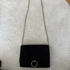 Svart mocka axelväska från H&M - Stilren svart axelväska i mocka från H&M med guldfärgad kedjerem och snygg ringdetalj framtill. Väskan har magnetlås och ljus insida med innerficka. Perfekt för dig som vill ha en enkel men trendig accessoar.