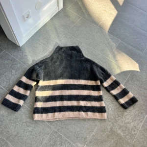 Randig stickad polotröja i svart och beige - Mysig stickad polotröja med breda svarta och beige ränder. Tröjan har en hög krage och är oversized med långa ärmar. Perfekt för kyliga dagar och passar snyggt till jeans eller kjol.