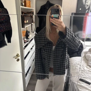 Rutig kappa med bälte och stora slag - Säljer en svart och grå rutig kappa med stora slag och bälte i midjan. Jackan har knappar framtill och ett klassiskt hundtandsmönster. Passformen är loose och den är perfekt för höst och vinter. Materialet känns som ullblandning.