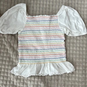 Vit blus med pastellränder Kappahl - Supersöt vit blus från Kappahl med kort puffärm och volangkant nertill. Framsidan har smock och tunna pastellfärgade ränder i rosa, blått, grönt, gult och orange. Perfekt för en färgglad och lekfull stil.