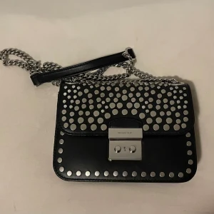 Svart axelväska Michael Kors med nitar - Snygg svart axelväska från Michael Kors med silverfärgade nitar i olika storlekar på locket och runt kanterna. Kedjerem i silver och lås framtill. 