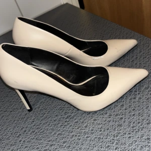 Beige pumps med spetsig tå från Zara - Stilrena pumps från Zara Basic i beige med spetsig tå och hög smal klack. Skorna har en klassisk siluett och är tillverkade i syntetmaterial med svart innersula. Perfekta för dig som gillar en elegant look med minimalistisk touch.