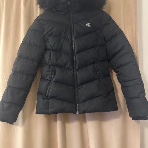 Svart pufferjacka från Calvin Klein - Snygg svart pufferjacka från Calvin Klein med huva och fuskpäls. Jackan har dragkedja framtill, två fickor med dragkedja och diskret CK-logo på bröstet. Perfekt för kalla dagar med sin vadderade design och stilrena look. Den är använd två gånger. Prisk kan diskuteras.💕