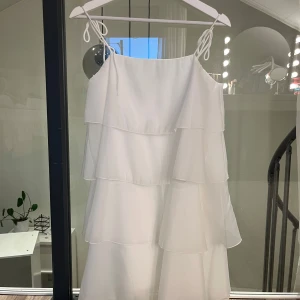 Vit volangklänning med knytband - Superfin vit klänning med flera lager volanger och tunna axelband som knyts upptill. Klänningen har en luftig och flowy känsla, perfekt för varma dagar. Materialet är lätt och ger en romantisk vibe.