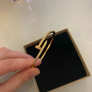 Guldarmband med glittriga stenar - Snyggt armband i guldton med unik design som liknar en böjd spik, dekorerat med små glittrande stenar längs ovansidan. Perfekt accessoar för att lyfta din outfit och ge en lyxig känsla. Kommer i en stilren ask.