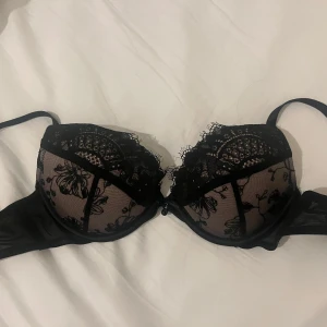 Svart spetsig push up BH - Säljer en svart bh med transparent mesh och vacker spets med blommigt mönster. Bh-n har en supersöt pärla i mitten. Den är storlek 70B och är från Hunkemöller