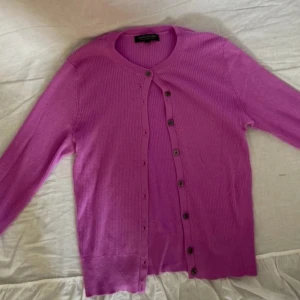 Lila kofta i merinoull från Banana Republic - Säljer en lila ribbstickad kofta från Banana Republic i 100% merinoull. Koftan har rund hals, knappar framtill och långa ärmar. Perfekt för lager-på-lager och ger en färgklick till din outfit.