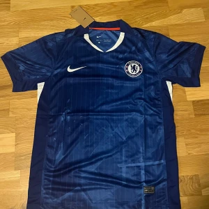 Chelsea FC hemmaställ 25/26 - Chelsea FCs officiella matchtröja i marinblått från Nike. Tröjan har korta ärmar, diskreta ränder och klubbmärke på bröstet. Tillverkad i lätt och ventilerande polyester med vita och röda detaljer på sidorna. 