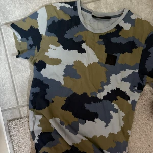 Camouflage t-shirt från Peak Performance - Säljer en kortärmad t-shirt från Peak Performance med snyggt camouflage-mönster i blått, grått, svart och olivgrönt. T-shirten har rund halsringning och en svart patch med logga på bröstet. Tillverkad i mjuk bomull som är skön att bära.