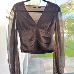 Brun blus med v-ringning från Bik Bok - Säljer en brun transparent blus från Bik Bok med v-ringning och långa, luftiga ärmar. Toppen har rynkade detaljer framtill och smockad nederkant för en snygg passform. Aldig använt köpt för 399kr. Skriv ifall du önskar mer bilder 🩷☺️