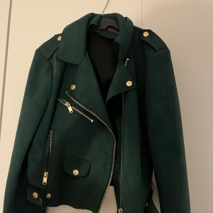 Mörkgrön bikerjacka från ONLY, stl 38 - Snygg mörkgrön bikerjacka från ONLY med guldiga dragkedjor och knappar. Jackan har klassisk krage, flera fickor med dragkedja och detaljer vid axlarna. Materialet är mjukt och mockaliknande, perfekt för dig som vill sticka ut.