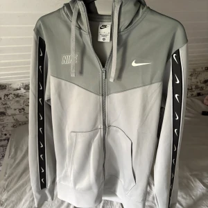 Grå Nike hoodie zip - Nike hoodie i två nyanser av grått med hel dragkedja och huva. Svarta detaljer med vita swoosh-loggor längs ärmarna. Praktiska fickor framtill och snörning i huvan. Storlek S
