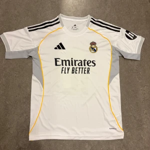 Real Madrid Mbappé 10  - Snygg vit Real Madrid fotbollströja från Adidas med Mbappé och nummer 10 på ryggen. Tröjan har svarta detaljer, gula linjer och grå partier på sidorna. Klassisk klubbmärke på bröstet och 'Emirates Fly Better' tryck. Tillverkad i lätt och ventilerande material.