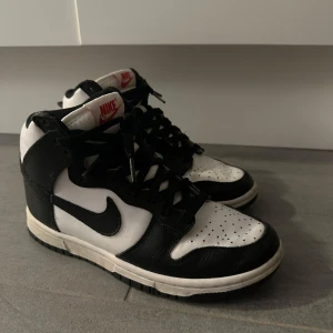 Nike Dunk High svart/vit - Svarta och vita Nike Dunk High med klassisk svart swoosh och röda Nike-loggor på plösen. Skorna har snörning, perforerad tå och är tillverkade i skinn. Perfekta för dig som gillar streetstyle och vill ha en ikonisk sneaker i garderoben. Säljer i samband med flytt därav priset!🤎