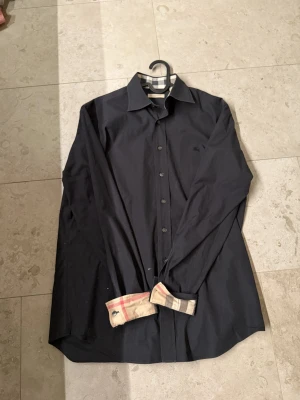 Mörkblå Burberry skjorta  - Säljer en mörkblå skjorta från Burberry med klassiska beige och svarta rutiga detaljer på insidan av kragen och manschetterna. Skjortan har lång ärm, knappar framtill. Perfekt för dig som gillar stilrena plagg med en twist.
