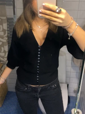 Svart v-ringad topp med knappar - Säljer en svart v-ringad topp med typ trekvarts ärmar och knappar hela vägen framtill nu. Modellen är croppad och har en clean, enkel look som passar till mycket. Materialet är mjukt och stretchigt, passar både till skola och fest.