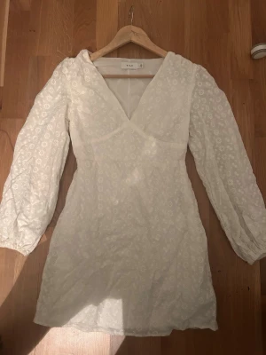 Vit kort klänning från Vila - Supersöt vit kort klänning från Vila med v-ringning och långa ärmar. Klänningen har broderade blommor över hela tyget och en lätt figurnära passform. Perfekt för sommarens alla tillfällen!