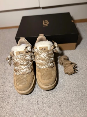 Uggs lowmel  - Säljer ett par snygga beige sneakers från UGG med breda, mönstrade snören i beige och vitt. Skorna har en chunky sula, detaljer i mocka och mesh samt fluffigt fårfoder på insidan för extra värme. Perfekta för dig som vill ha både stil och komfort.