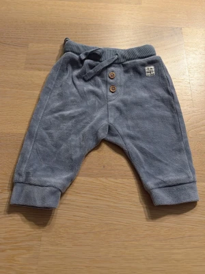 Blå mjukisbyxor från Lindex baby - Supersköna blå mjukisbyxor från Lindex för nyfödd. Byxorna har ribbad midja med snörning, två dekorativa knappar framtill och mjuka muddar vid bensluten. Tillverkade i ett mjukt material som känns bekvämt mot huden.