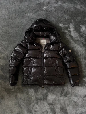 Svart Moncler pufferjacka med huva - Säljer en svart pufferjacka från Moncler med glansig finish och huva. Jackan har dragkedja framtill, två fickor med dragkedja och en ärmficka med Moncler-logga. Insidan har ett coolt serietryck och snörning i nederkant. Perfekt för kalla dagar.
