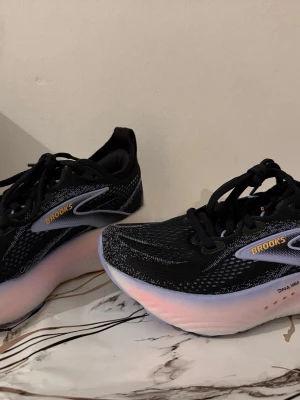 Nya Oanvända Brooks Glycerin 22 Dam svarta sneakers med rosa sula - Säljer ett par snygga Brooks sneakers i svart mesh med vita och grå detaljer samt logga i gult. Skorna har en tjock, vit och rosa sula och snörning framtill. Perfekta för dig som gillar sportig stil och vill ha bekväma skor till träning eller vardag.
