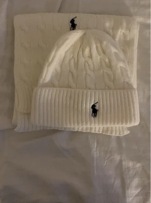Vit kabelstickad mössa & halsduk Polo Ralph Lauren - Säljer ett set med vit kabelstickad mössa och matchande halsduk från Polo Ralph Lauren. Båda har den klassiska loggan broderad i svart och är gjorda i mjuk ull. Perfekt för kalla dagar och riktigt snyggt till vinterlooken.
