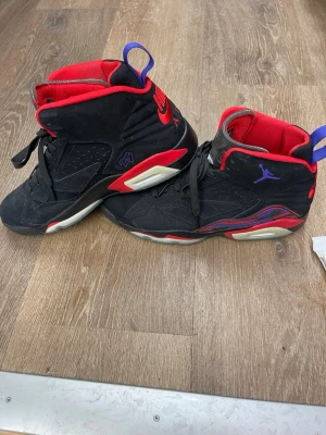 Nike Air Jordan 6 Retro svart/röd - Säljer ett par Nike Air Jordan 6 Retro sneakers i svart mocka med röda och lila detaljer. Skorna har hög siluett, snörning och en ikonisk Jumpman-logga på sidan och sulan. Insidan är fodrad i rött tyg och yttersulan har genomskinliga partier.