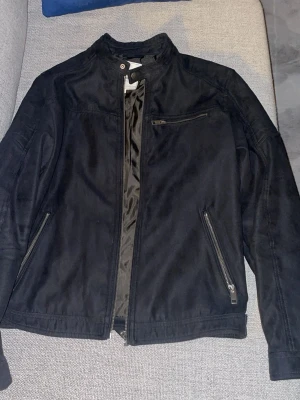 Svart mocka jacka med dragkedjor - Snygg mörkblå jacka med ståkrage och flera dragkedjor, inklusive två snedställda fickor och en bröstficka. Jackan har en stilren look och passar perfekt till både jeans och sneakers. Fodrad insida för extra komfort.