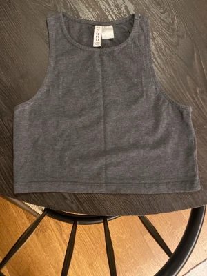 Mörkgrå croppad linnetopp H&M - Säljer en enkel och stilren mörkgrå croppad linnetopp från H&M Basic. Toppen är ärmlös med hög halsringning. Materialet är mjukt och stretchigt, vilket gör den superbekväm.