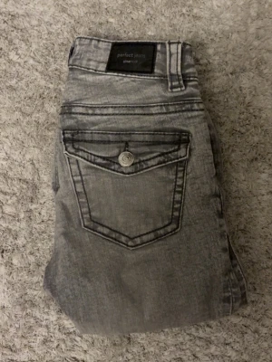 Grå bootcut jeans från Gina tricot Perfect Jeans - Säljer ett par grå jeans från Gina tricot Perfect Jeans i storlek 32. De har en snygg bootcut-modell, klassiska fem fickor och coola detaljer på bakfickan med knapp. Jeansen har en tvättad look och är tillverkade i ett mjukt bomullsmaterial.