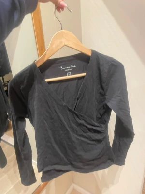 Svart omlottopp från Bondelid - Säljer en svart omlottopp från Bondelid i storlek S. Toppen har lång ärm, v-ringning och omlottdesign som ger en snygg siluett. Tillverkad i mjuk bomull som känns skön mot huden. Perfekt att styla med jeans eller kjol.