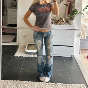 Lågmidjade slitna bootcut jeans - Säljer ett par ljusblå bootcut jeans med låg midja och slitna detaljer på benen. Jeansen har klassisk femficksdesign och är tillverkade i denim. Perfekta för dig som gillar en avslappnad och trendig look.