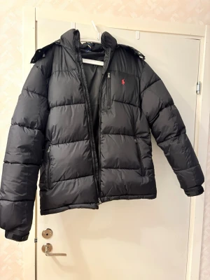 Svart pufferjacka från Polo Ralph Lauren - Säljer en svart pufferjacka från Polo Ralph Lauren med huva och dragkedja. Jackan har en röd broderad logga på bröstet och två sidofickor med dragkedja. Perfekt för kalla dagar och riktigt skön att bära.