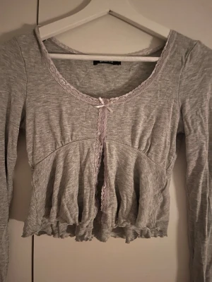 Grå croppad kofta med spets från Gina Tricot - Supersöt grå croppad kofta från Gina Tricot med spetsdetaljer längs kanterna och en liten rosett framtill. Långärmad och med volangkant nertill, perfekt för lager på lager och en söt touch till din outfit.