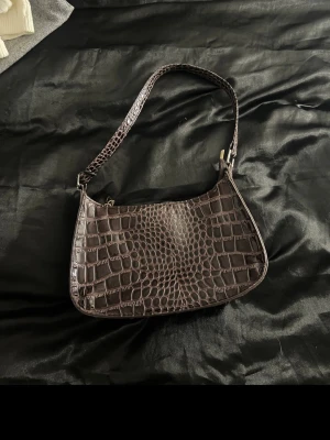 Brun axelväska i krokopräglat skinn - Säljer en brun axelväska med croco-mönster och stilren form. Väskan har ett kort axelband och är gjord i syntetmaterial som liknar skinn. Perfekt för dig som gillar trendiga accessoarer med lite edge.