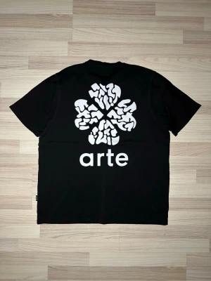 Svart t-shirt från Arte med tryck - Svart t-shirt från Arte med stor vit grafisk print och logga på ryggen. Klassisk passform med rund hals och korta ärmar. Tillverkad i mjuk bomull som känns skön mot huden. Perfekt för dig som gillar stilrena plagg med en twist.