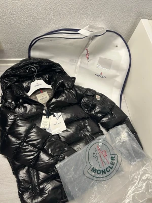Svart Moncler Maya pufferjacka med huva, följer med galjärn och dustbag - Säljer en svart glansig pufferjacka från Moncler med huva och dragkedja. Jackan har Moncler-logga på ärmen, praktiska fickor och quiltad design. Insidan är grå och jackan är fylld med dun för extra värme. Perfekt för kalla dagar och riktigt snygg streetstil. Det finns en ficka på insidan av jackan. NFC tag funkar. Huvan kan man ta av. Allt ni ser på bilden följer med, galgarna följer med samt dustbag. Överiga frågor eller om ni vill få mer bilder kontakta gärna mig ;) ( storlek M) (kan sänkas)