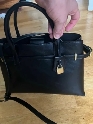 MNG svart handväska - Svart handväska med handtag och axelrem. Väskan har en liten ficka med dragkedja inuti och en dekorativ detalj med ett litet hänglås.