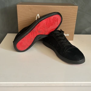 Svarta sneakers från Christian Louboutin - Snygga svarta sneakers från Christian Louboutin i mocka med klassisk röd sula och svart snörning. Skorna har rund tå och en stilren design som sticker ut med den ikoniska röda undersidan. Perfekta för dig som vill ha något exklusivt och trendigt.