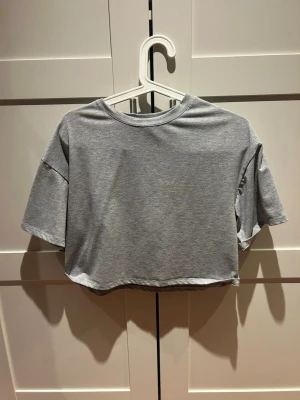 Grå croppad t-shirt - En enkel och stilren grå croppad t-shirt med korta ärmar och rund hals. Tillverkad i mjuk bomull som känns skön mot huden. Perfekt till jeans eller shorts för en avslappnad look.