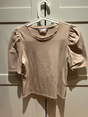 Beige topp med puffärm från ONLY - Säljer en beige topp från ONLY med puffiga korta ärmar och rund halsringning. Toppen är i mjuk bomull och har en enkel, stilren design som passar till mycket. Perfekt för dig som gillar en clean look med en twist.