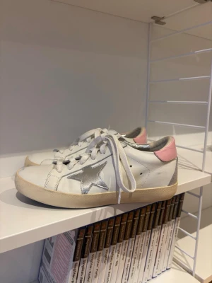Golden Goose skor - Säljer ett par vita sneakers i skinn med silverfärgad stjärna på sidan och rosa detalj vid hälen. Skorna har snörning och en beige sula. Insidan är metallic-silver och de har en klassisk låg profil. Perfekta för dig som gillar en clean men ändå lekfull look.
