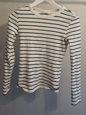 Randig långärmad topp Gina Tricot - Säljer en vit långärmad topp från Gina Tricot med svarta horisontella ränder. Modellen är enkel och klassisk med rund halsringning och smal passform. Perfekt basplagg till garderoben och lätt att matcha med olika stilar.