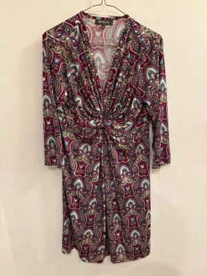 Mönstrad kortklänning från Alice Bizous - Kortklänning med paisleyinspirerat mönster i vinrött, blått, beige och svart. Djup v-ringning och twistad detalj i midjan som ger en snygg siluett. Trekvartsärm och mjukt, stretchigt material som känns skönt mot huden.
