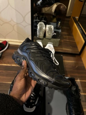 Svarta Nike Air Max Plus sneakers - Säljer ett par helsvarta Nike Air Max Plus med ikonisk vågig design och synliga luftbubblor i sulan. Skorna har snörning, rund tå och är tillverkade i syntetmaterial med gummisula. Perfekta för dig som vill ha en clean och sportig look.
